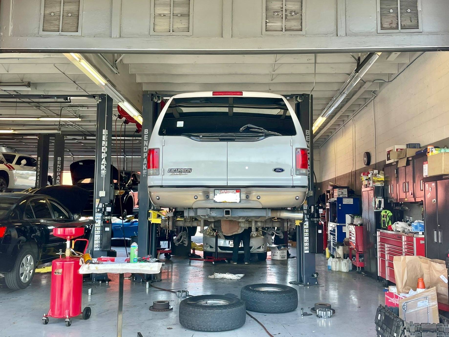 Inside a vehicle repair shop, cars on lifts, tools, American flag, ceiling with skylights, open garage door.